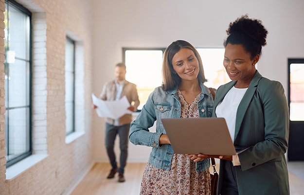 2 Frauen schauen auf einen Laptop, lächeln und stehen in einer Wohnung. Im Hintergrund steht ein Mann mit Wohnungsskizze.